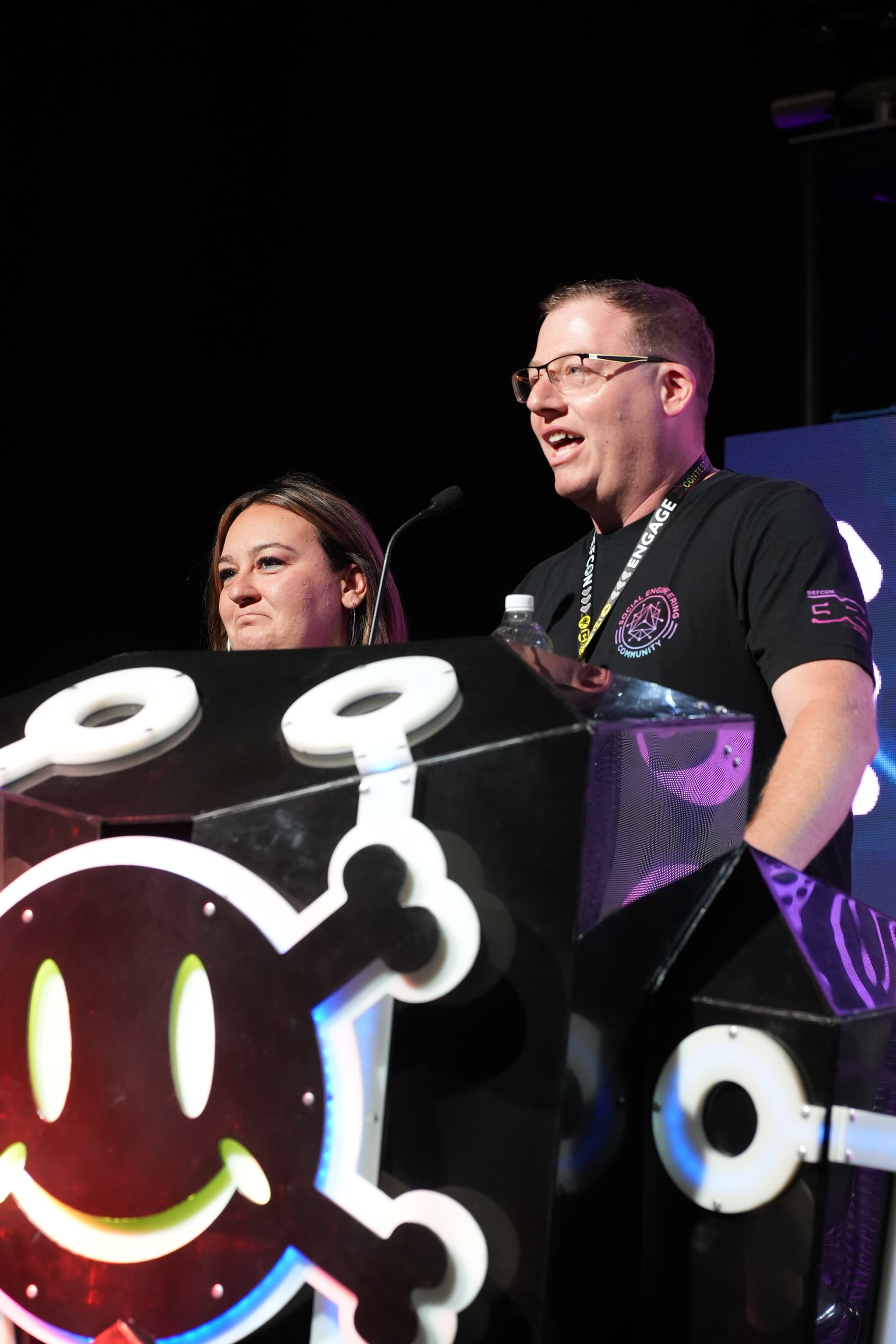 Two people standing behind a podium decorated with a smiling skull and crossbones logo, speaking into microphones