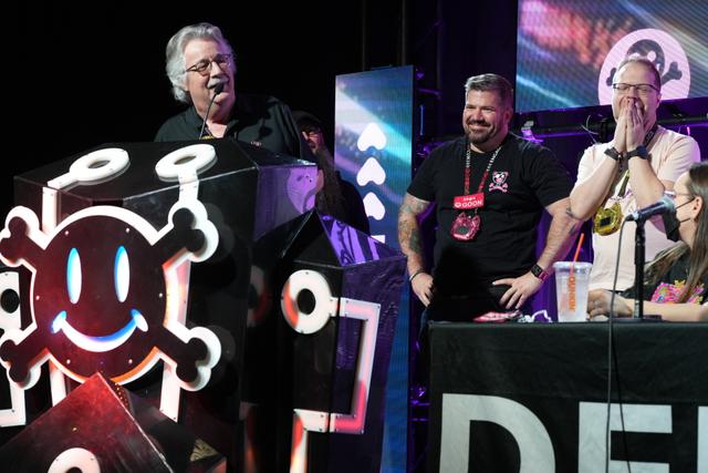 A group of people on a stage, with one person speaking at a podium decorated with a smiling skull and crossbones symbol. Two other individuals stand nearby, one with a hand over their mouth, and another person is seated at a table with the word DEF visible. The background features colorful lighting and screens