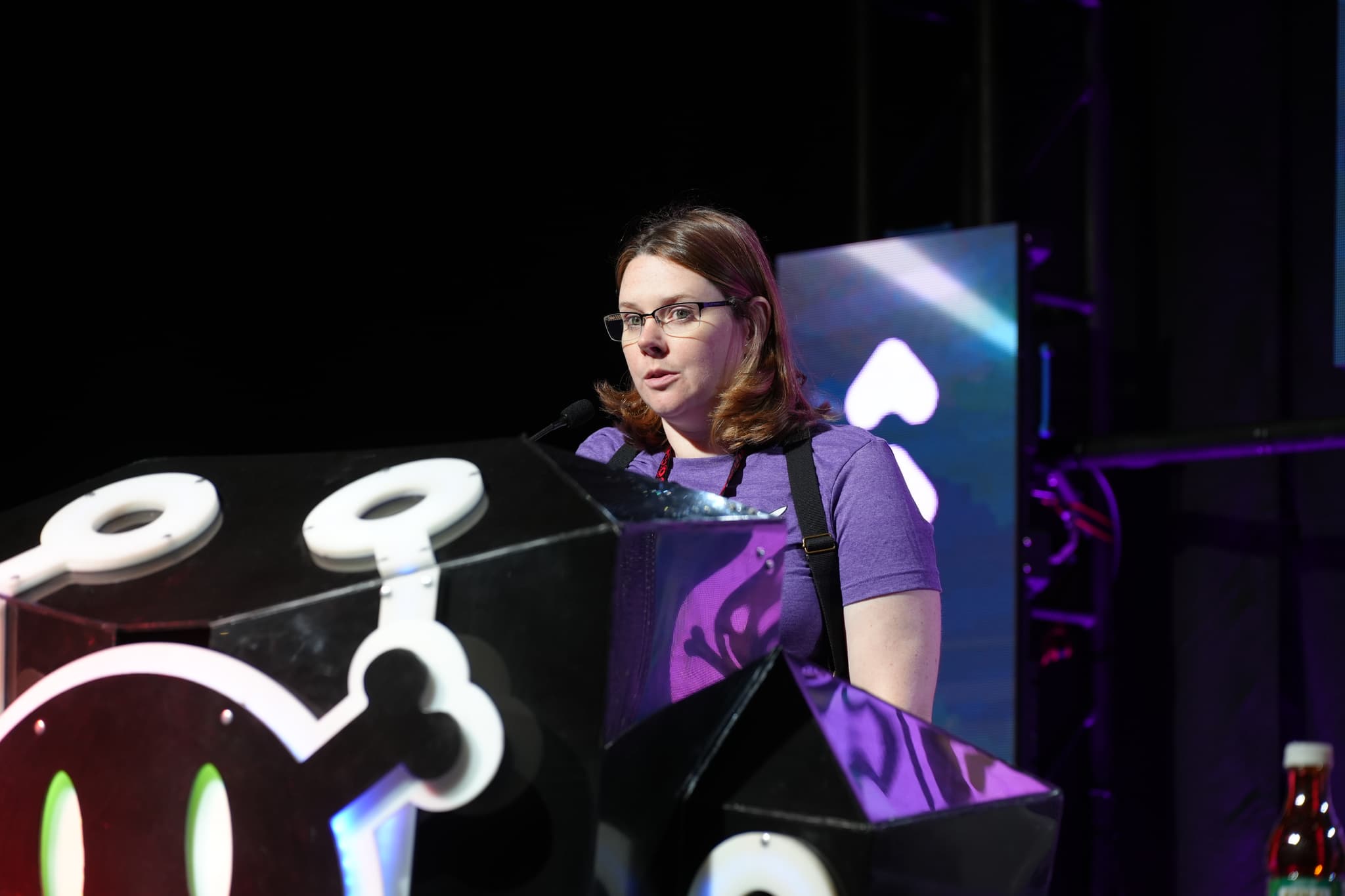 A person standing behind a podium, speaking into a microphone, with a dark background and colorful lighting