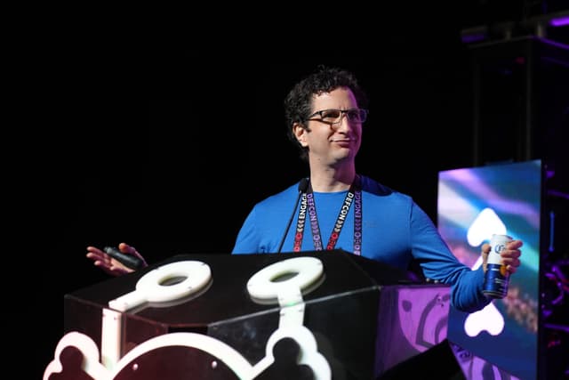 A person wearing glasses and a blue shirt stands behind a large, decorated podium, holding a microphone and a small object, with a dark background and colorful lights