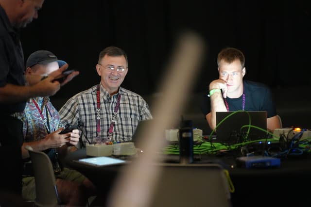 Several people sitting at a table with laptops and electronic devices, engaged in conversation and activities, in a dimly lit environment