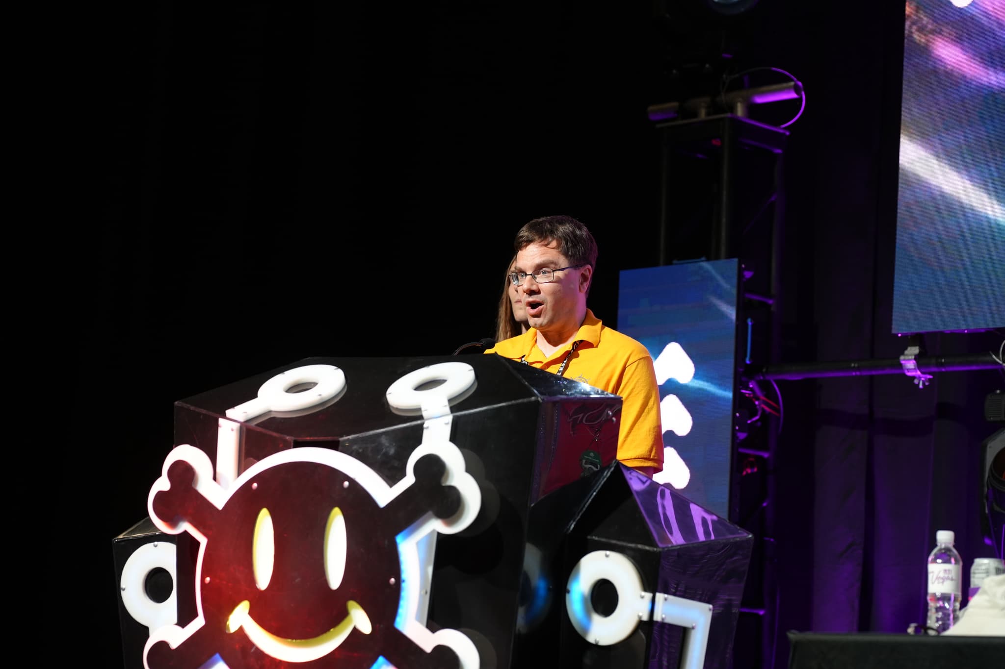 A person standing behind a podium with a large, colorful emblem, speaking or presenting on a stage with a dark background