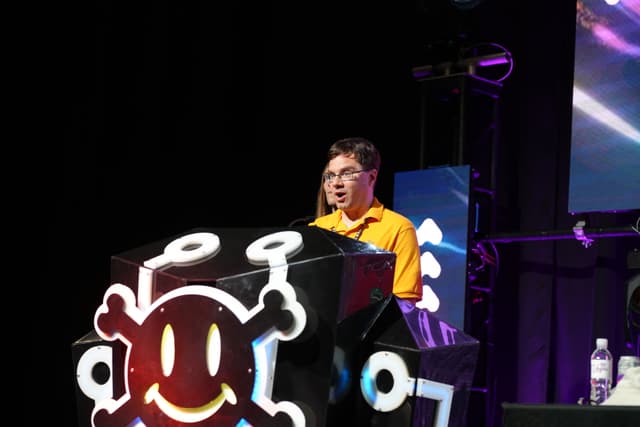 A person standing behind a podium with a large, colorful emblem, speaking or presenting on a stage with a dark background