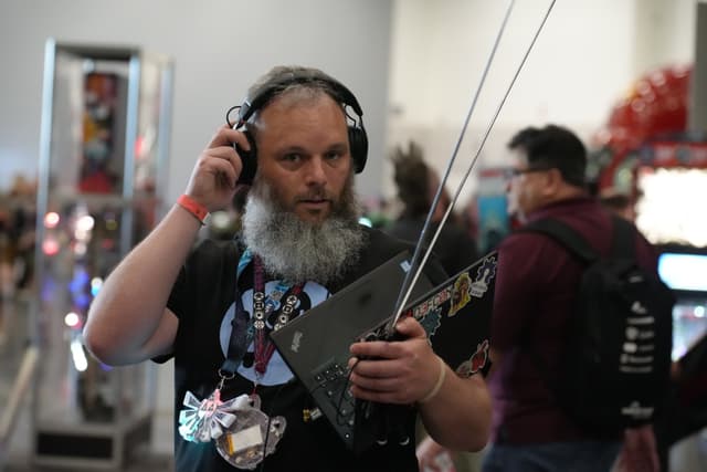 A bearded man wearing headphones and holding a laptop with antennas, standing in a busy indoor setting with other people in the background