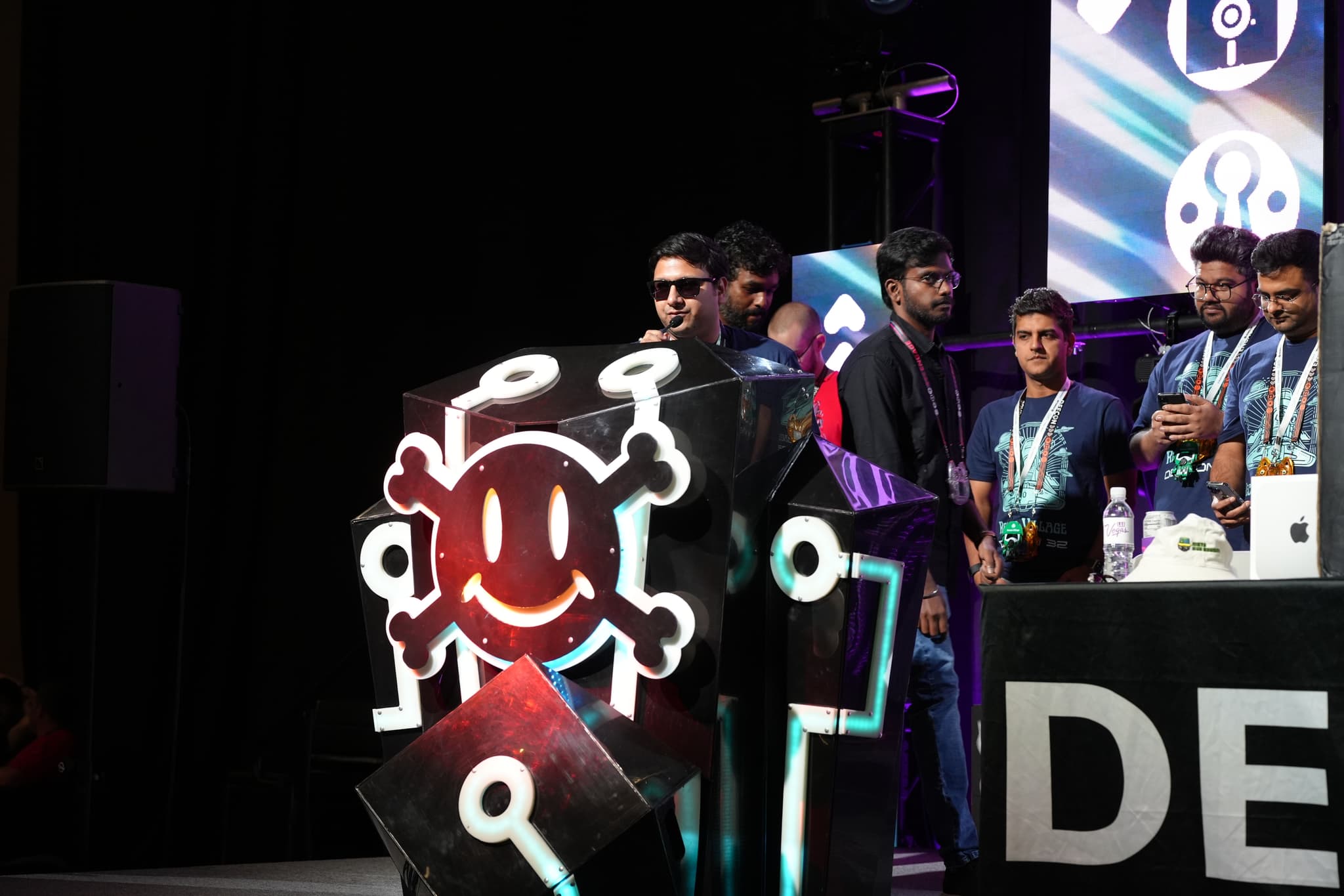 A group of people standing on a stage, with one person speaking at a podium decorated with a smiling skull and crossbones symbol. The background features a large screen with various symbols, and several individuals are wearing lanyards