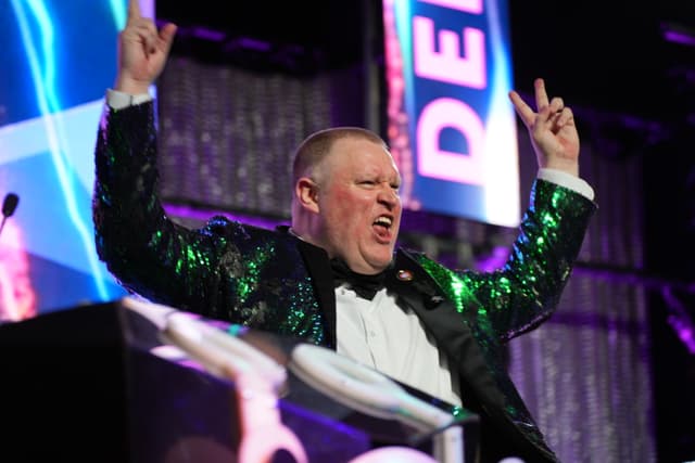 A man in a sparkly green jacket enthusiastically gestures with both hands raised, standing behind a podium with colorful lights and signs in the background