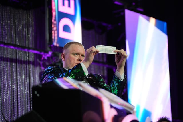 A person holding up a piece of paper on a stage with colorful lighting and a shimmering backdrop