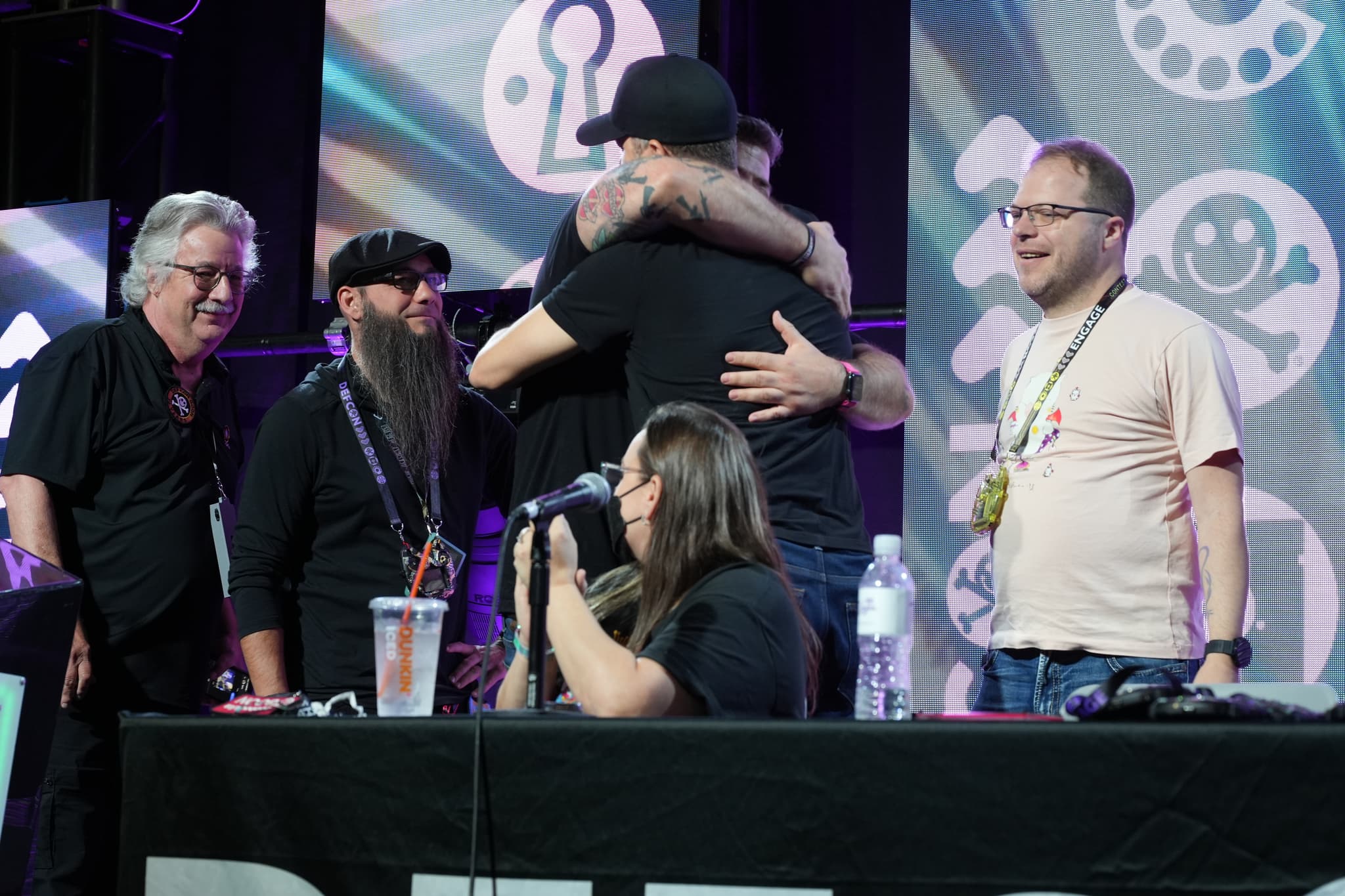 A group of people are gathered on a stage, with two individuals hugging in the center. A woman is seated at a table with a microphone, and others are standing around, some smiling. The background features colorful, patterned designs