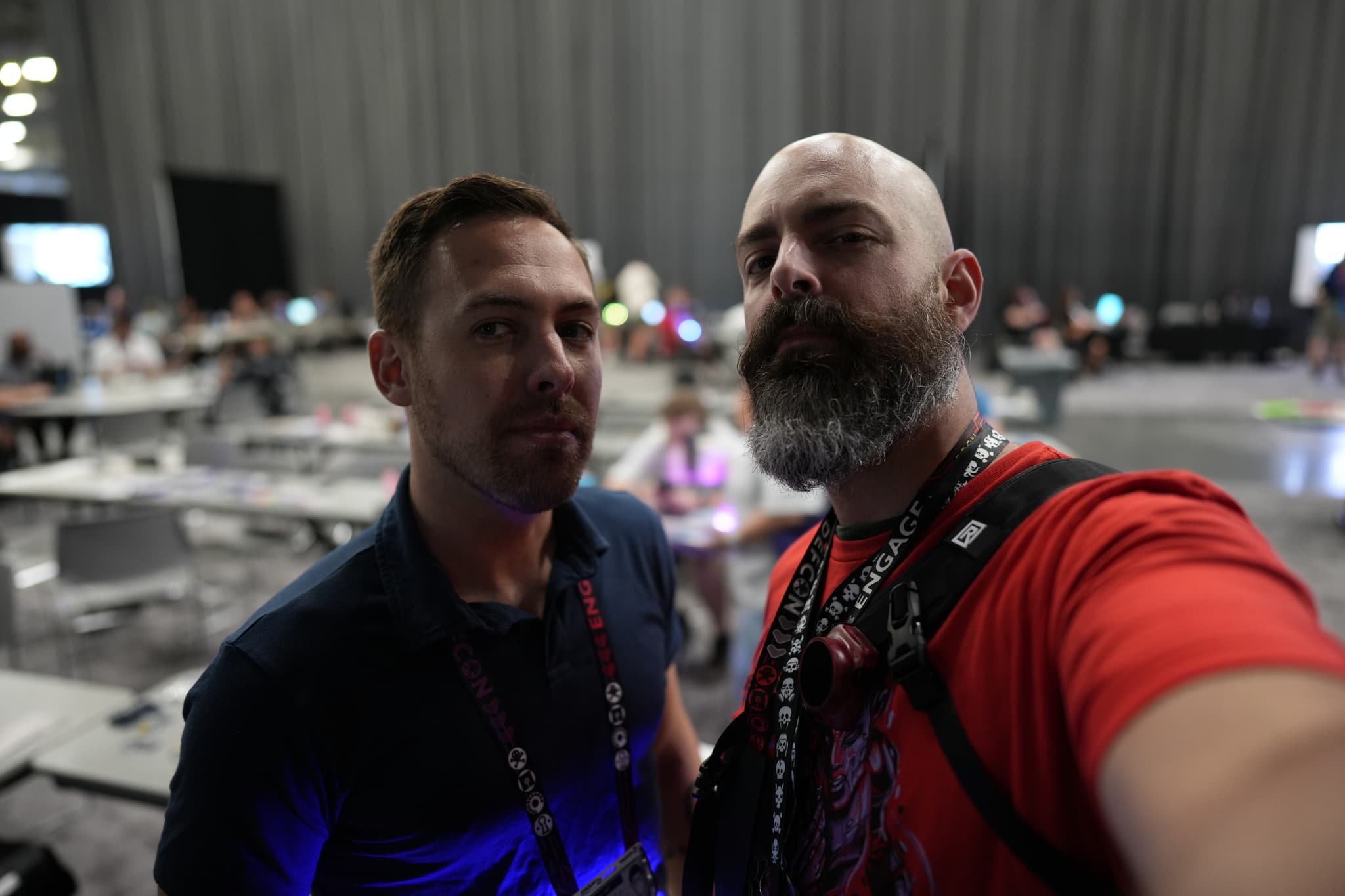 Two men taking a selfie in a large indoor space with tables and people in the background