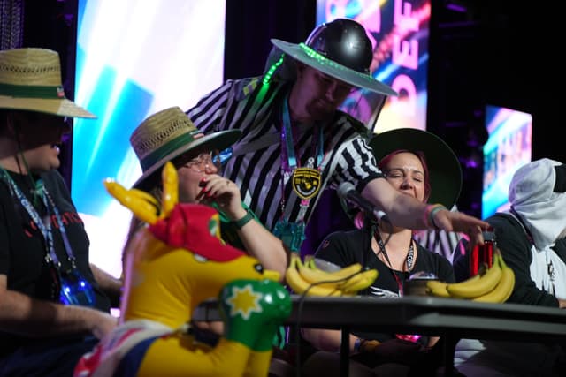 A group of people wearing hats and costumes are seated at a table with inflatable toys and bananas, with a colorful, illuminated background behind them