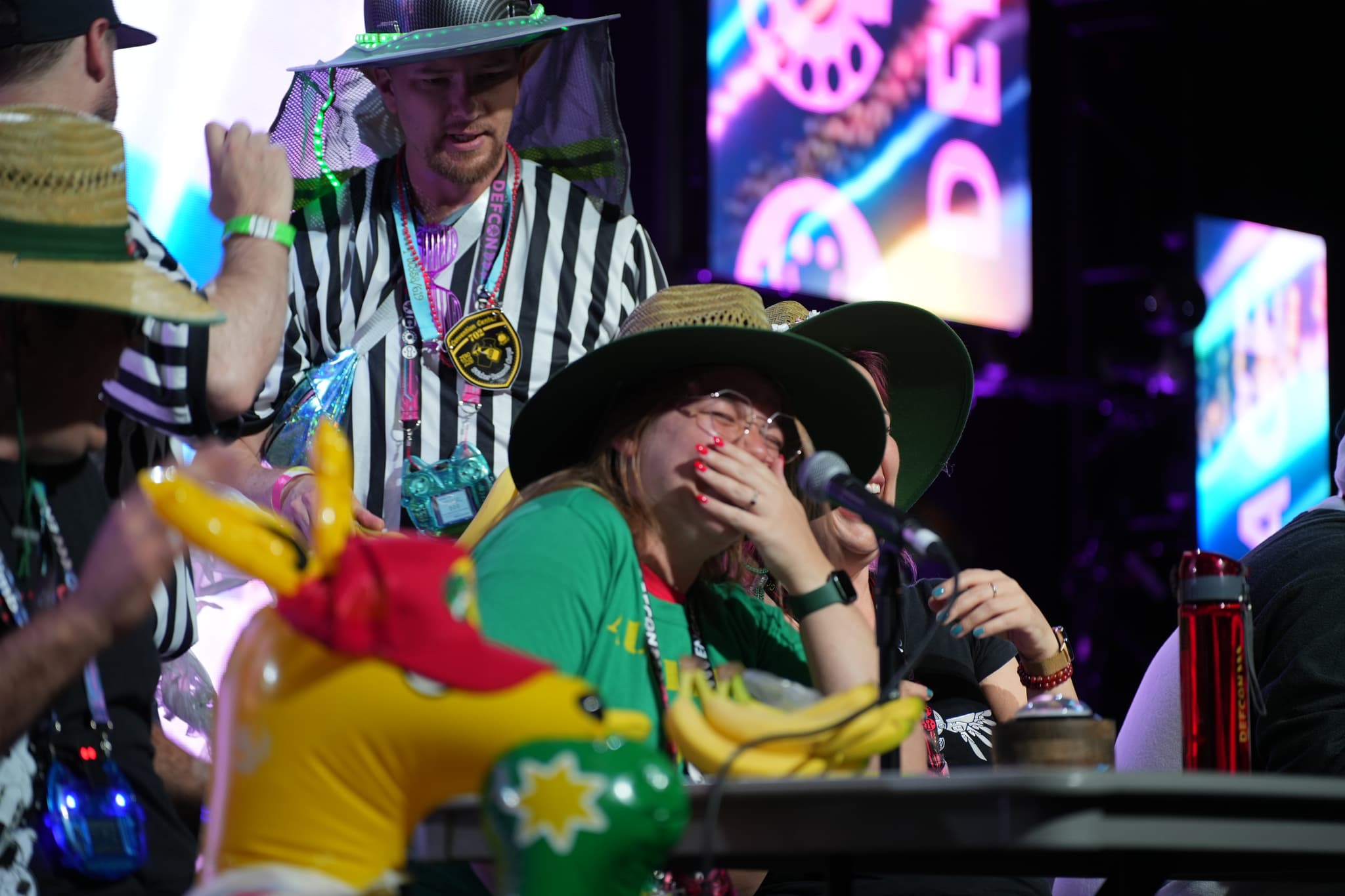 A group of people wearing hats and colorful clothing, with one person covering their mouth in surprise or excitement. The background features bright lights and screens, and there is a yellow inflatable object in the foreground