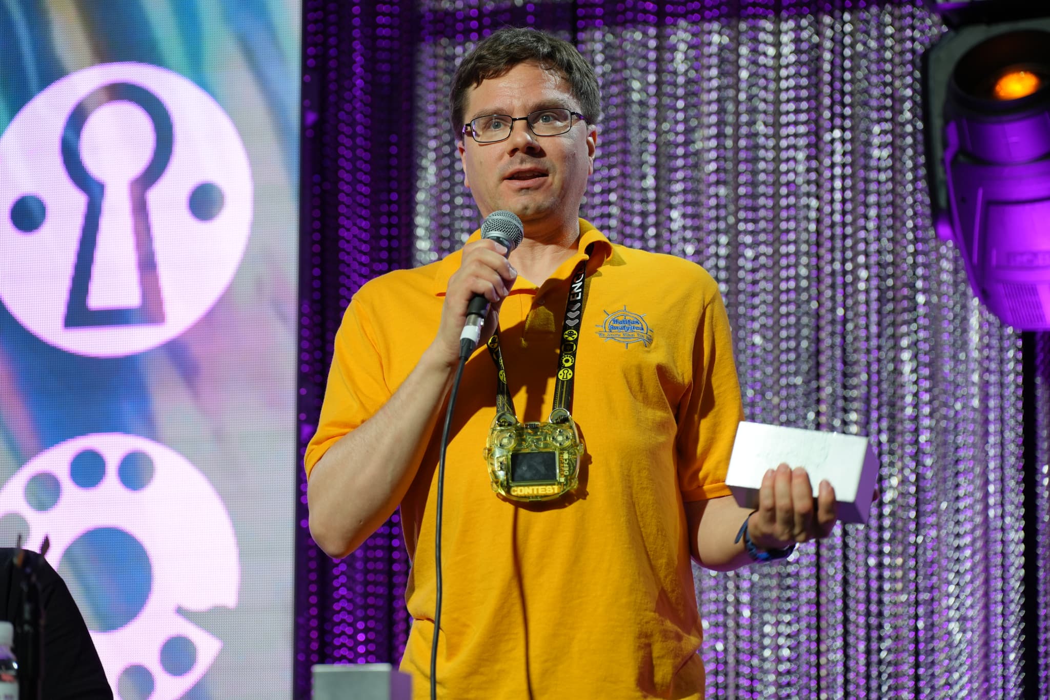 A man in a yellow shirt is speaking into a microphone on a stage, holding a piece of paper. The background features a sparkly curtain and a large screen with various symbols