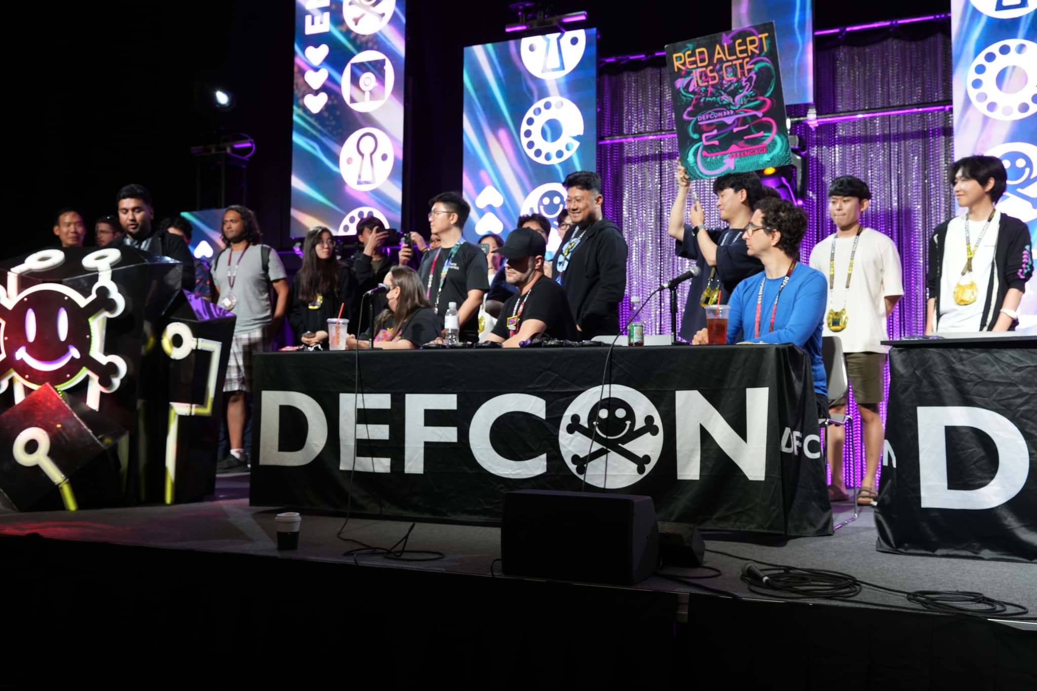 A group of people standing behind tables with DEFCON banners, on a stage with colorful lighting and digital displays in the background