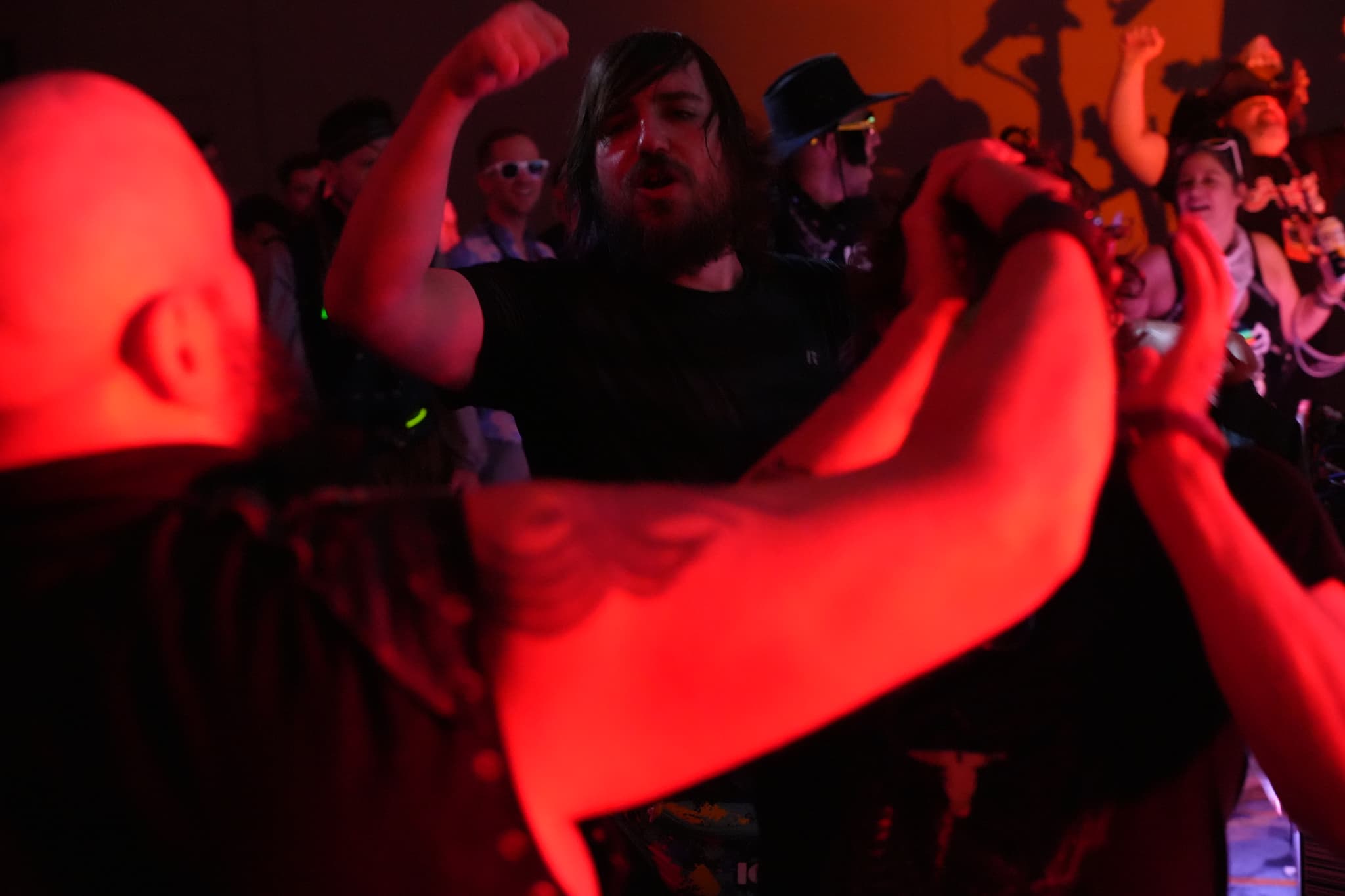 A group of people at a concert or event, with one person in the foreground flexing their arm and others in the background cheering and enjoying the atmosphere. The lighting is predominantly red