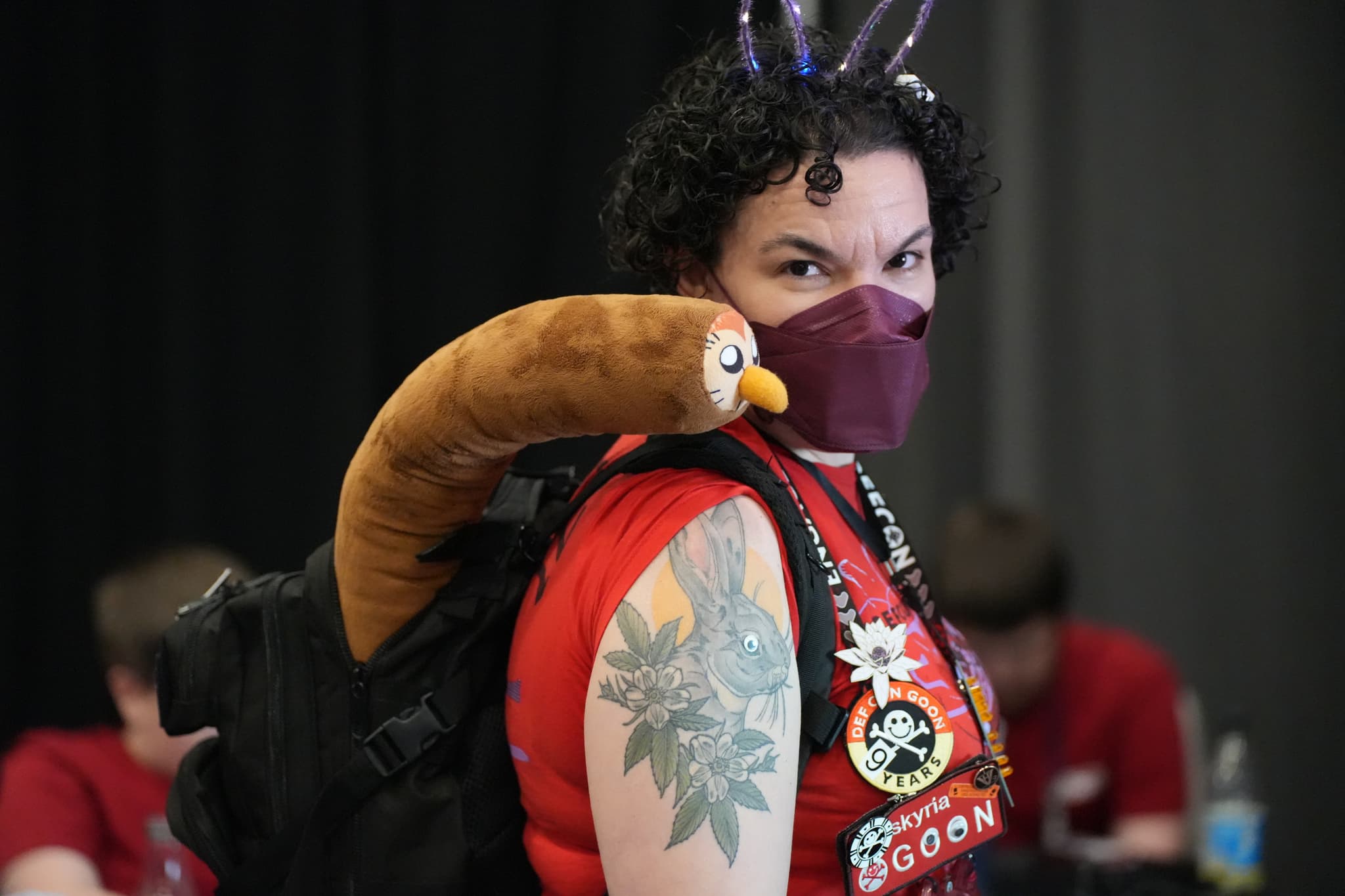 A person wearing a red shirt, a purple face mask, and a backpack with a plush toy resembling a sausage with a face. The person has curly hair, a tattoo on their arm, and is adorned with various pins and badges