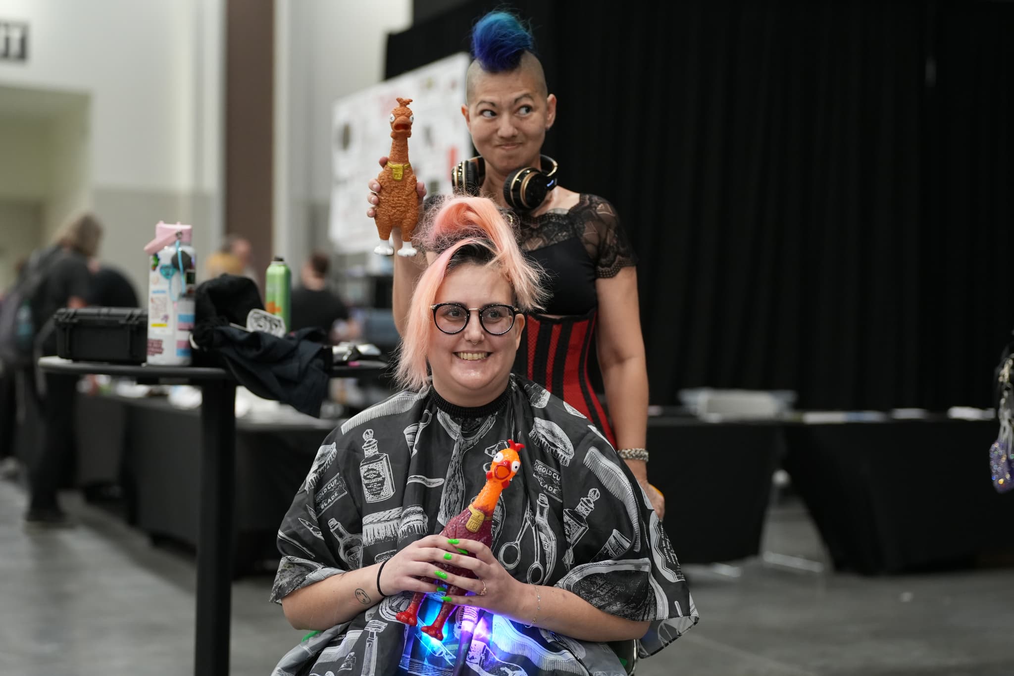 Two individuals in a salon setting, one seated with a colorful toy and the other standing behind with a unique hairstyle. The background includes salon equipment and other people