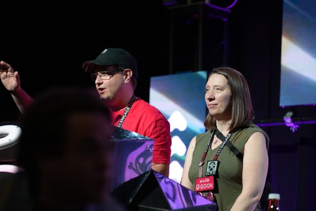 Two people standing in front of a stage with colorful lighting, one wearing a red shirt and black cap, and the other wearing a green top