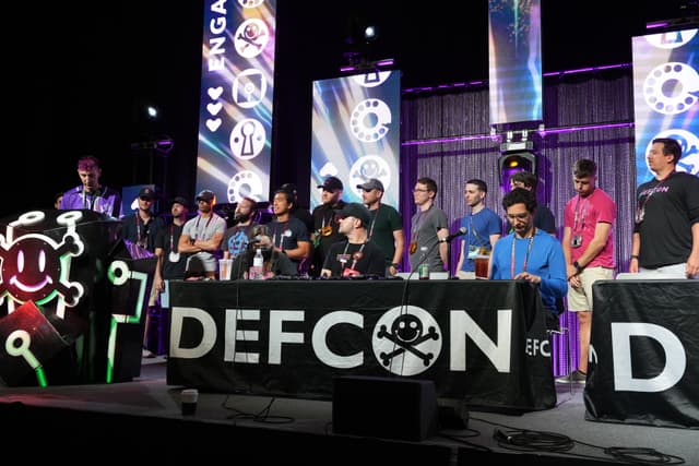 A group of people standing and sitting behind a table with a DEFCON banner, featuring a skull and crossbones logo, on a stage with colorful lighting and digital displays in the background