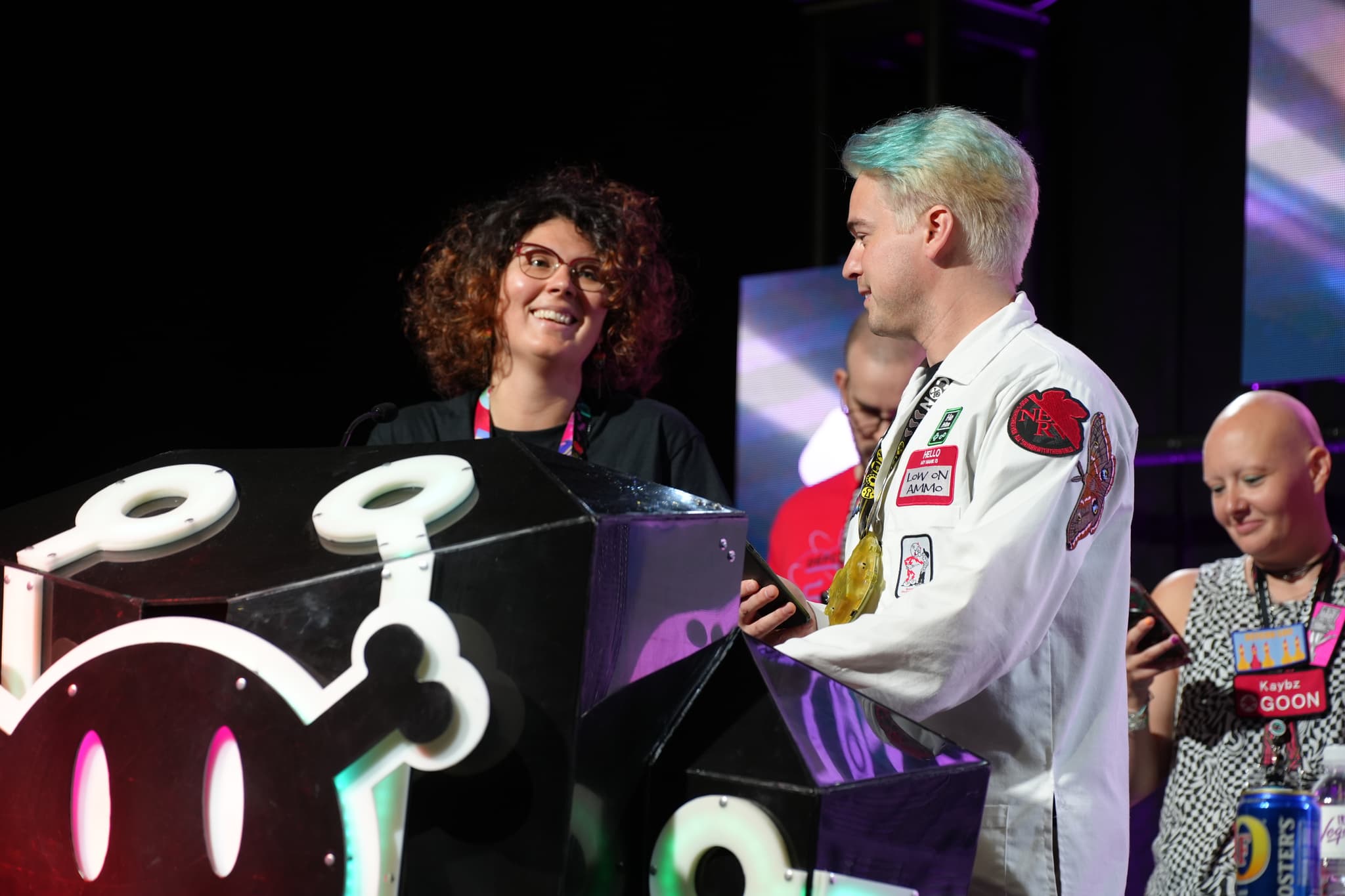 A group of people standing on a stage, engaging with a large, decorated console. One person has curly hair and is smiling, while another has light-colored hair with a blue tint and is wearing a jacket with patches. Two other individuals are in the background, one of whom is bald and also smiling