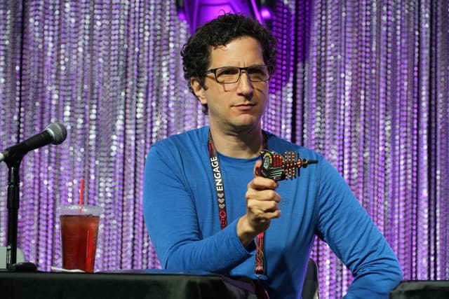 A man in a blue shirt holding a toy gun, sitting at a table with a microphone and a drink, against a backdrop of shimmering curtains