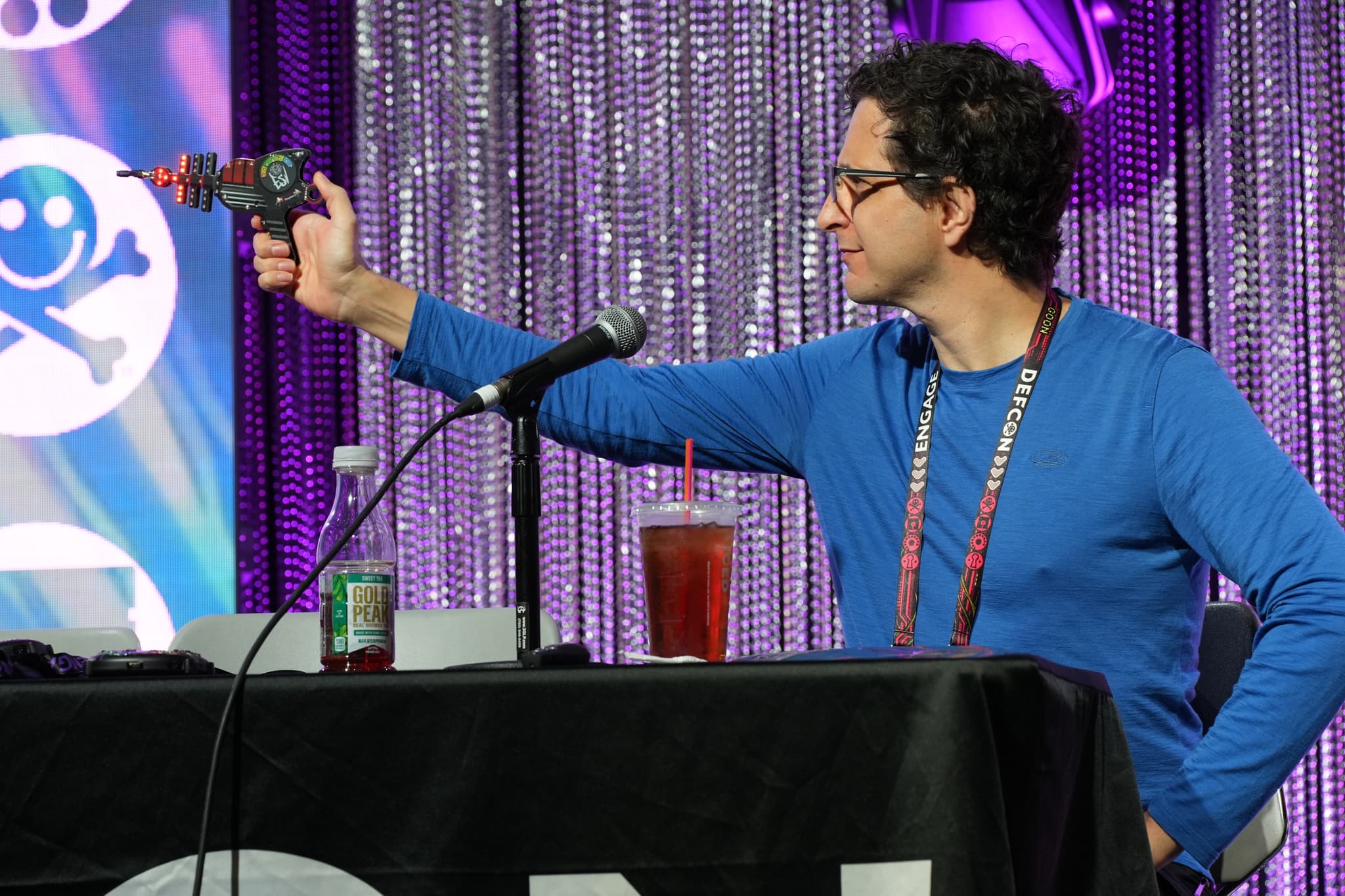 A person in a blue shirt is sitting at a table with a microphone, holding a toy ray gun. The background features colorful decorations and a drink is placed on the table