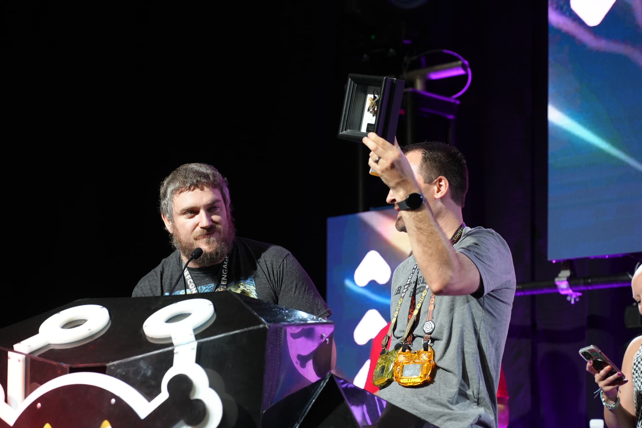 Two individuals on a stage, one holding up a trophy or award, with a large screen and a decorated podium in the background