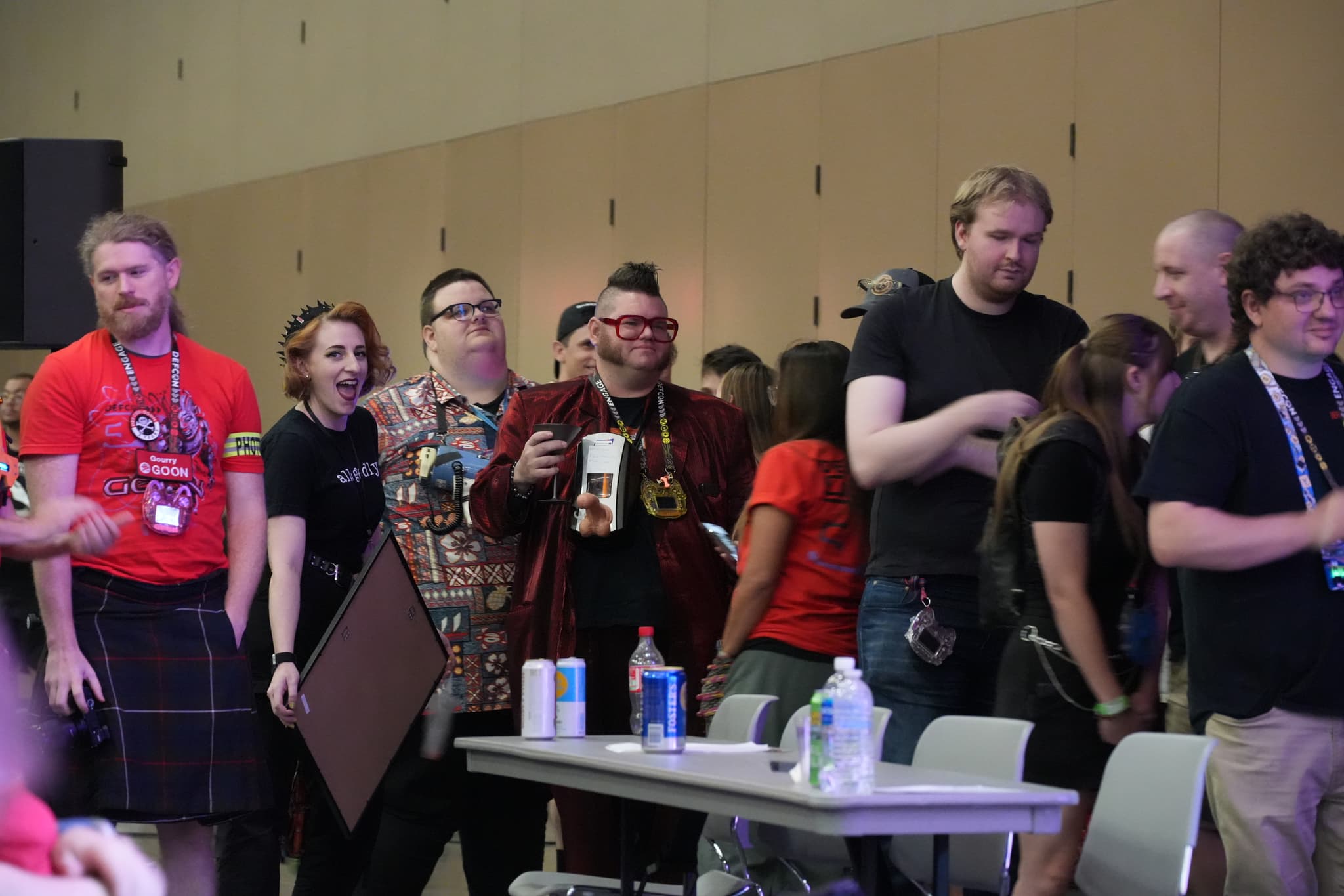 A group of people standing and interacting in a room, some wearing lanyards and badges, with a table in the foreground holding drinks and other items