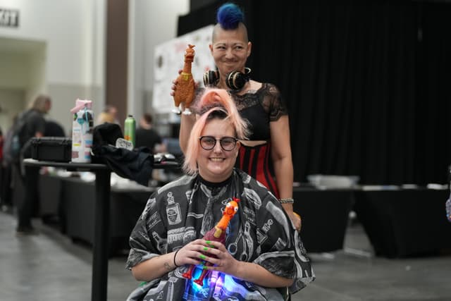 Two people with unique hairstyles, one seated and one standing behind, both holding rubber chickens, in an indoor setting with a black curtain in the background