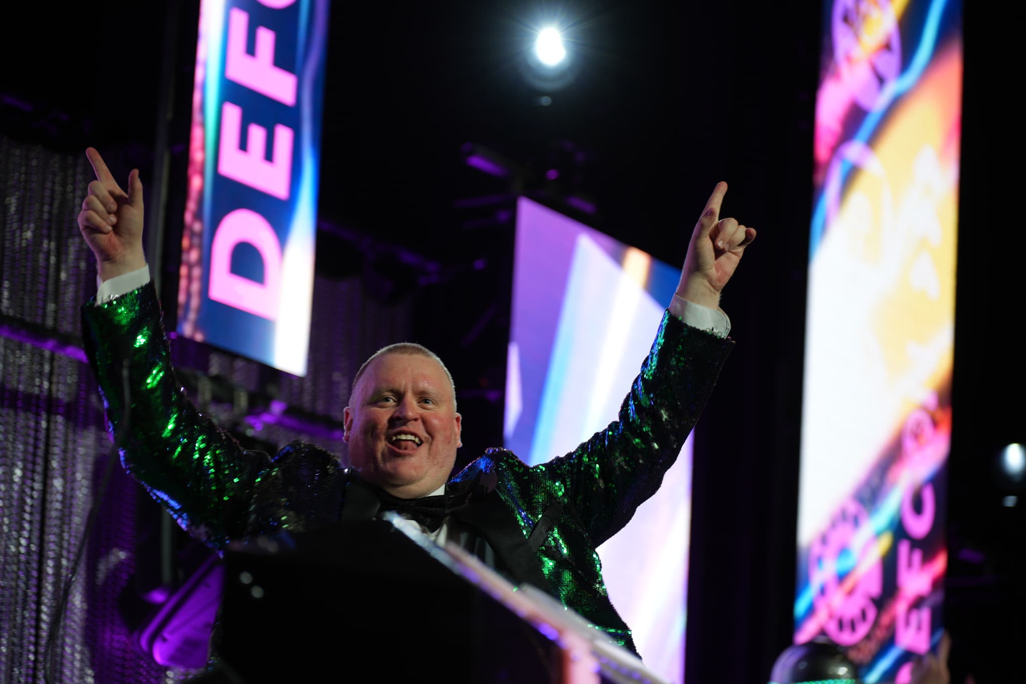 A person in a shiny green jacket is raising both arms with index fingers pointing upwards, surrounded by colorful, illuminated signs