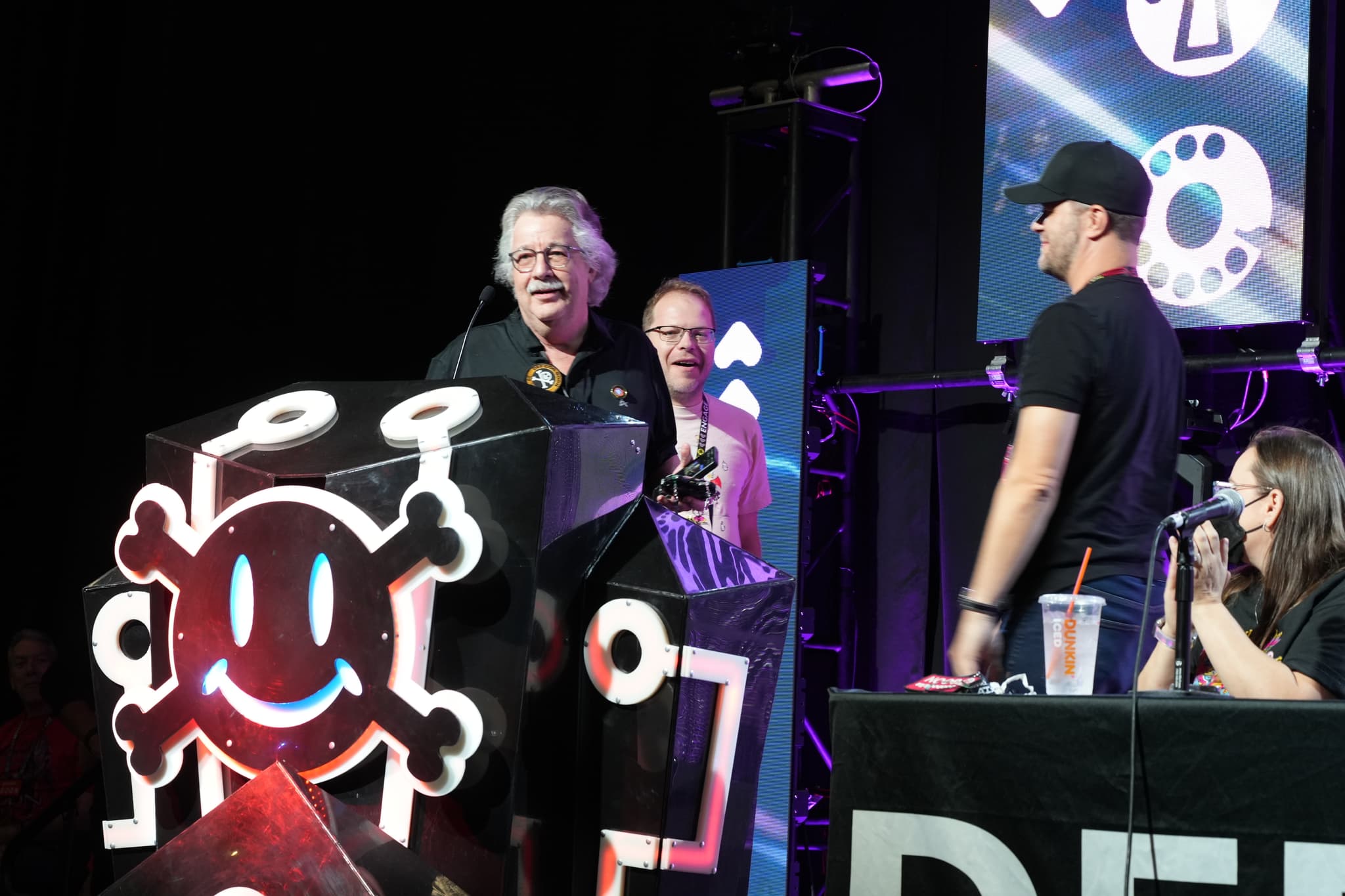 A group of people on a stage, with one person speaking at a podium decorated with a smiling skull and crossbones design. Other individuals are standing and sitting nearby, with various electronic and decorative elements in the background