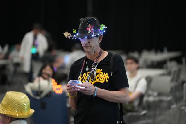 A person wearing a hat adorned with lights and decorations, looking at a device in their hands, with other people and tables in the background