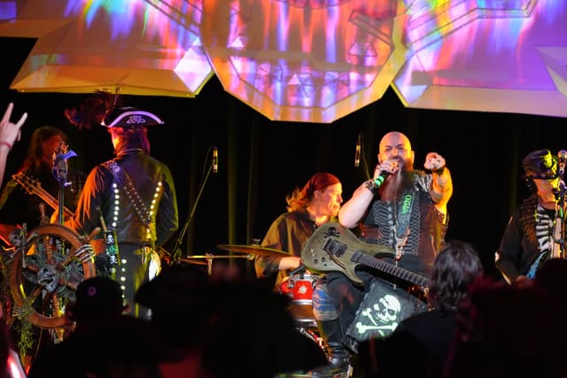 A band performing on stage with colorful lighting effects, featuring a lead singer, guitarist, drummer, and other musicians dressed in elaborate costumes