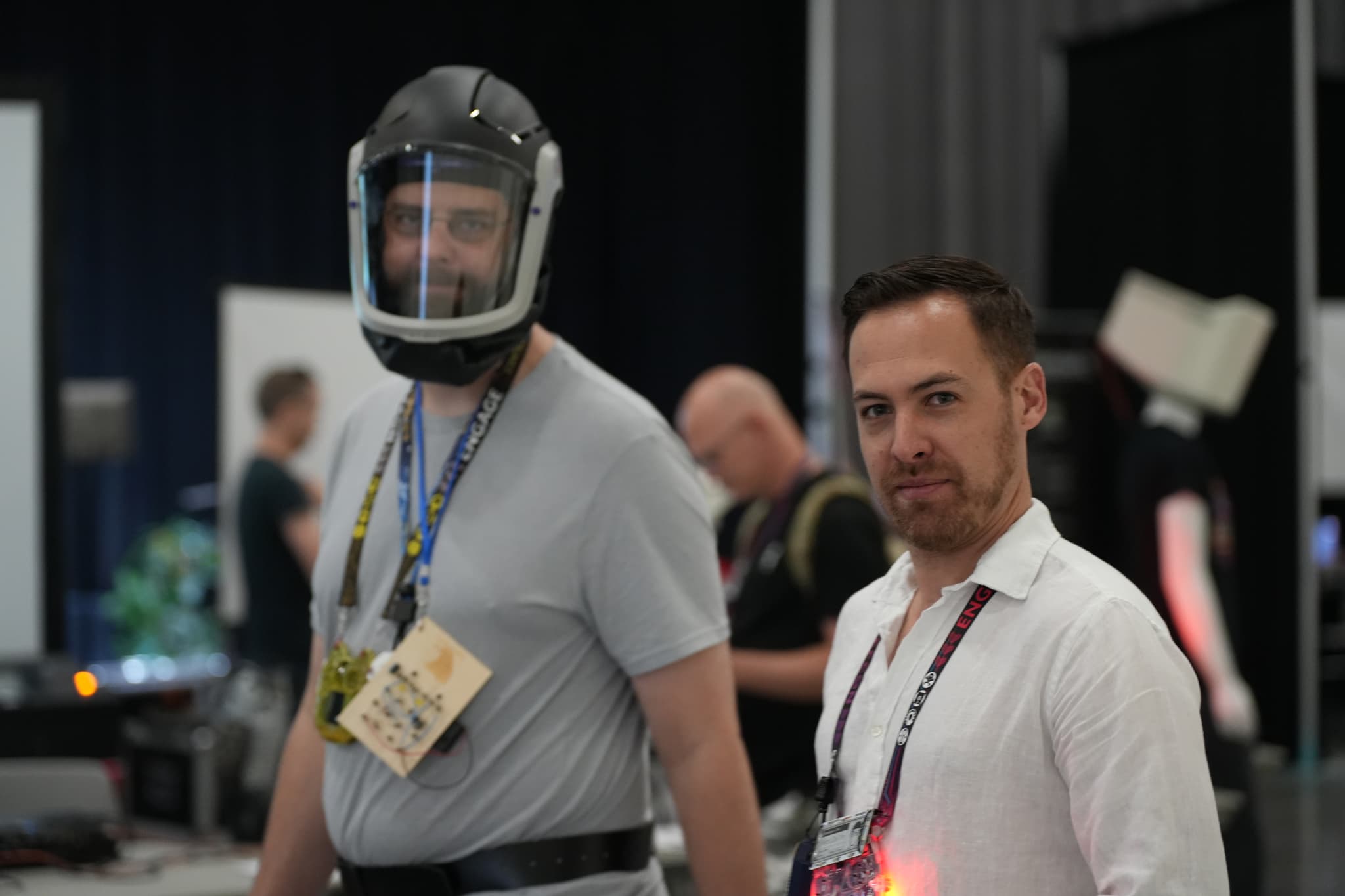 Two men standing indoors, one wearing a helmet and a gray shirt with a badge around his neck, and the other in a white shirt with a lanyard. Several people and equipment are visible in the background