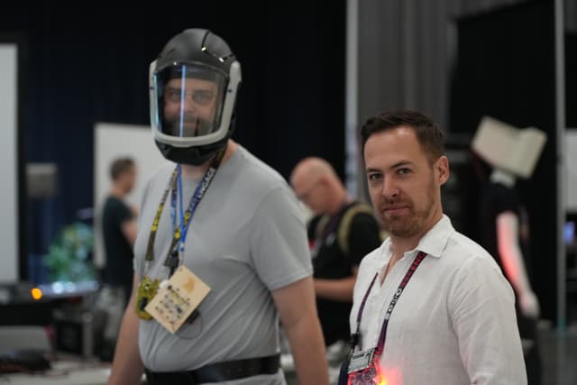 Two men standing indoors, one wearing a helmet and a gray shirt with a badge around his neck, and the other in a white shirt with a lanyard. Several people and equipment are visible in the background