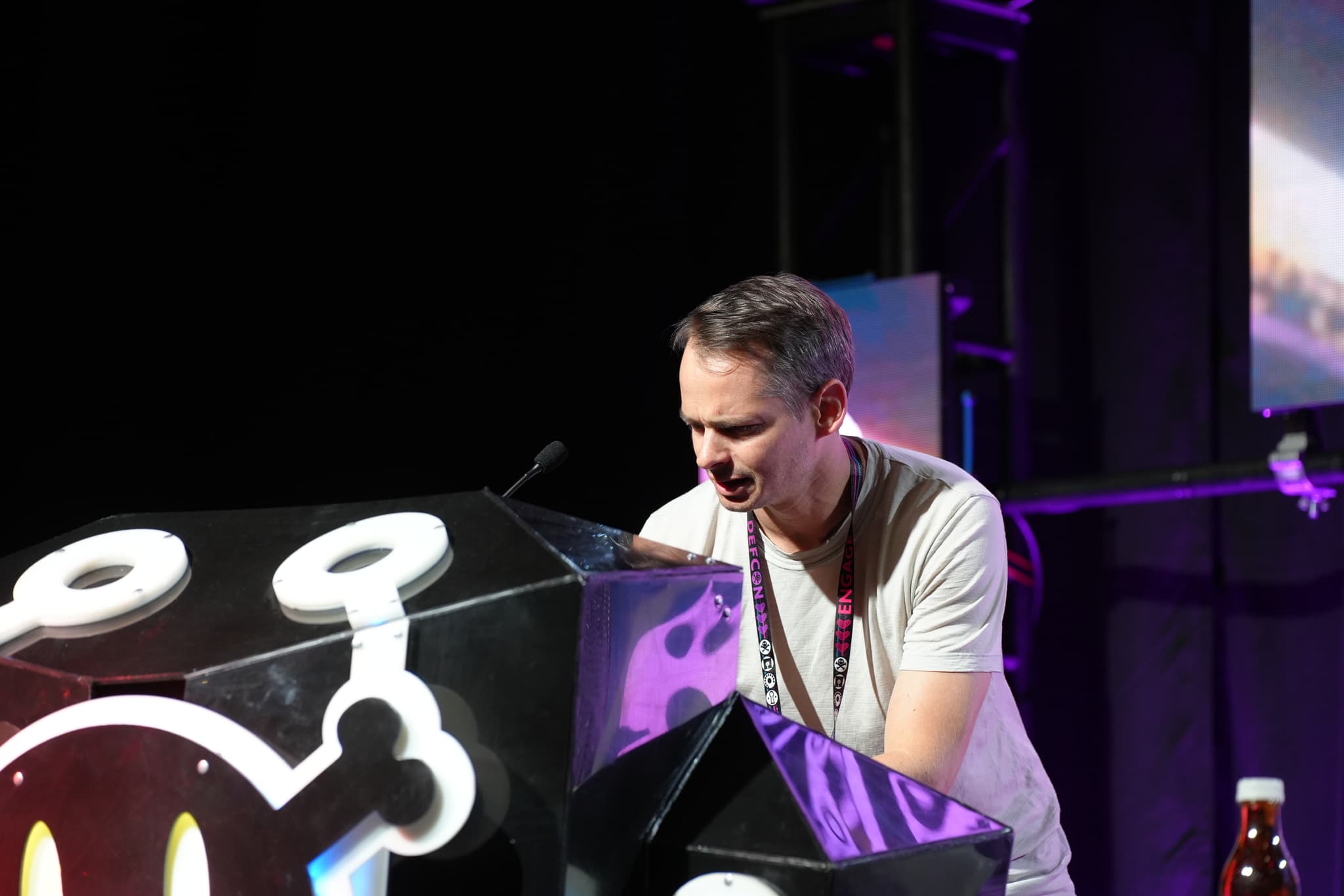 A man is standing behind a podium decorated with a cartoonish bomb design, speaking or presenting on a stage with a dark background and some lighting equipment visible