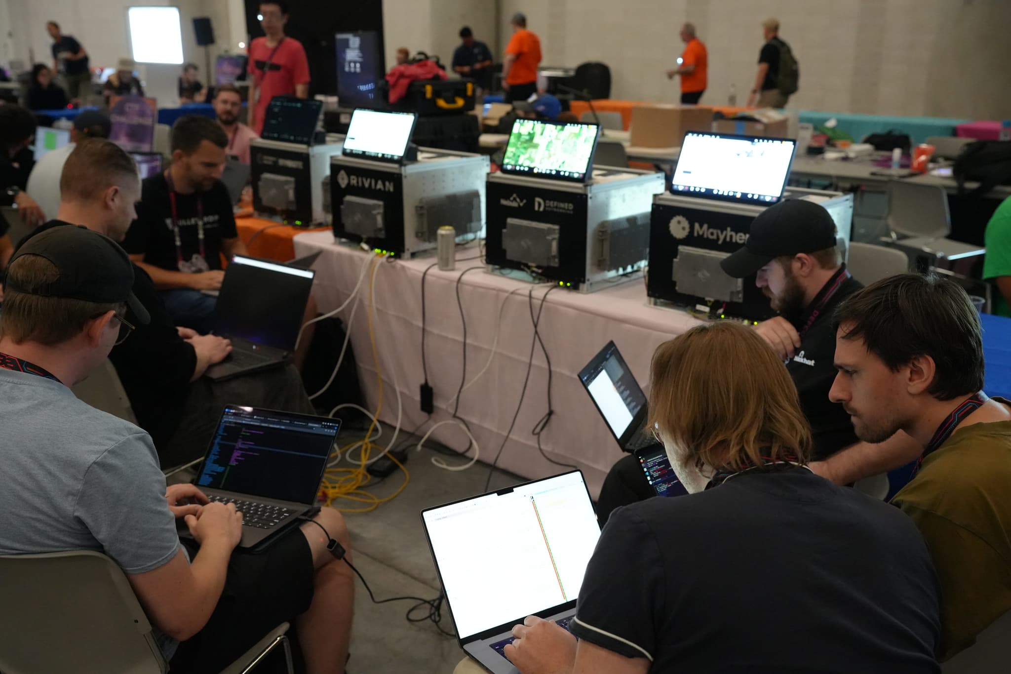 A group of people working on laptops in a room with multiple computer setups and screens, with others in the background