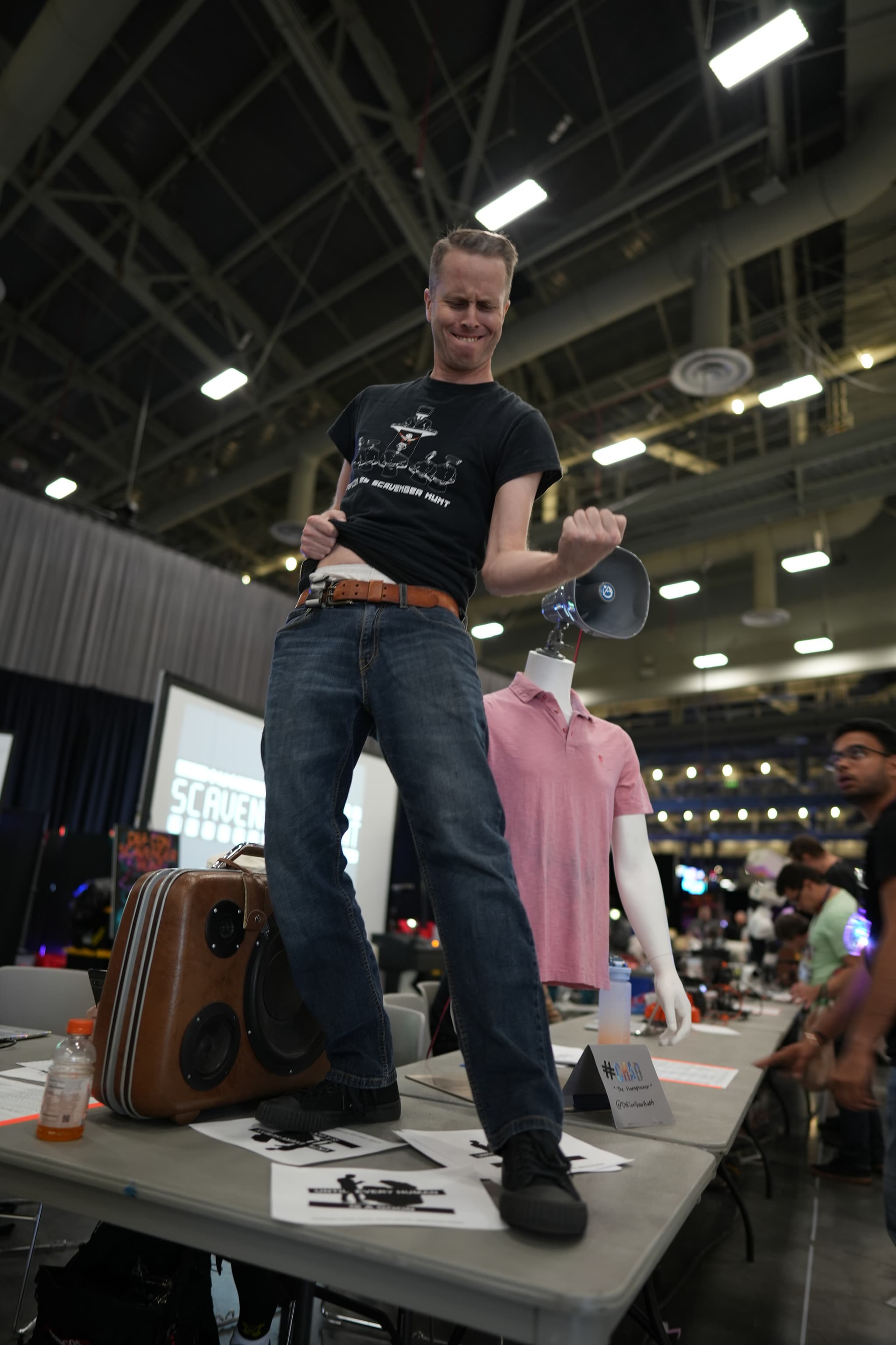 A person standing on a table, holding a megaphone, and striking a dynamic pose in a large indoor venue with various items and people in the background