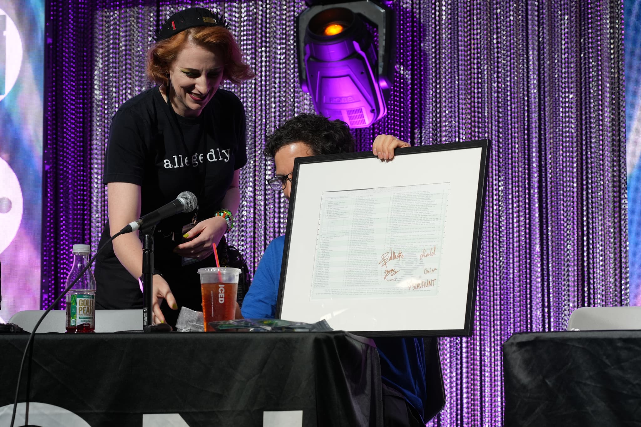 Two people at a table, one holding a framed document and the other reaching for a microphone, with a purple curtain backdrop and stage lighting