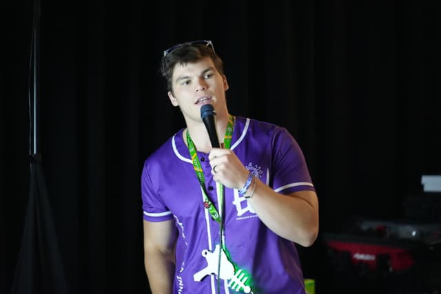 A person wearing a purple shirt and a lanyard, holding a microphone and speaking, with a dark background