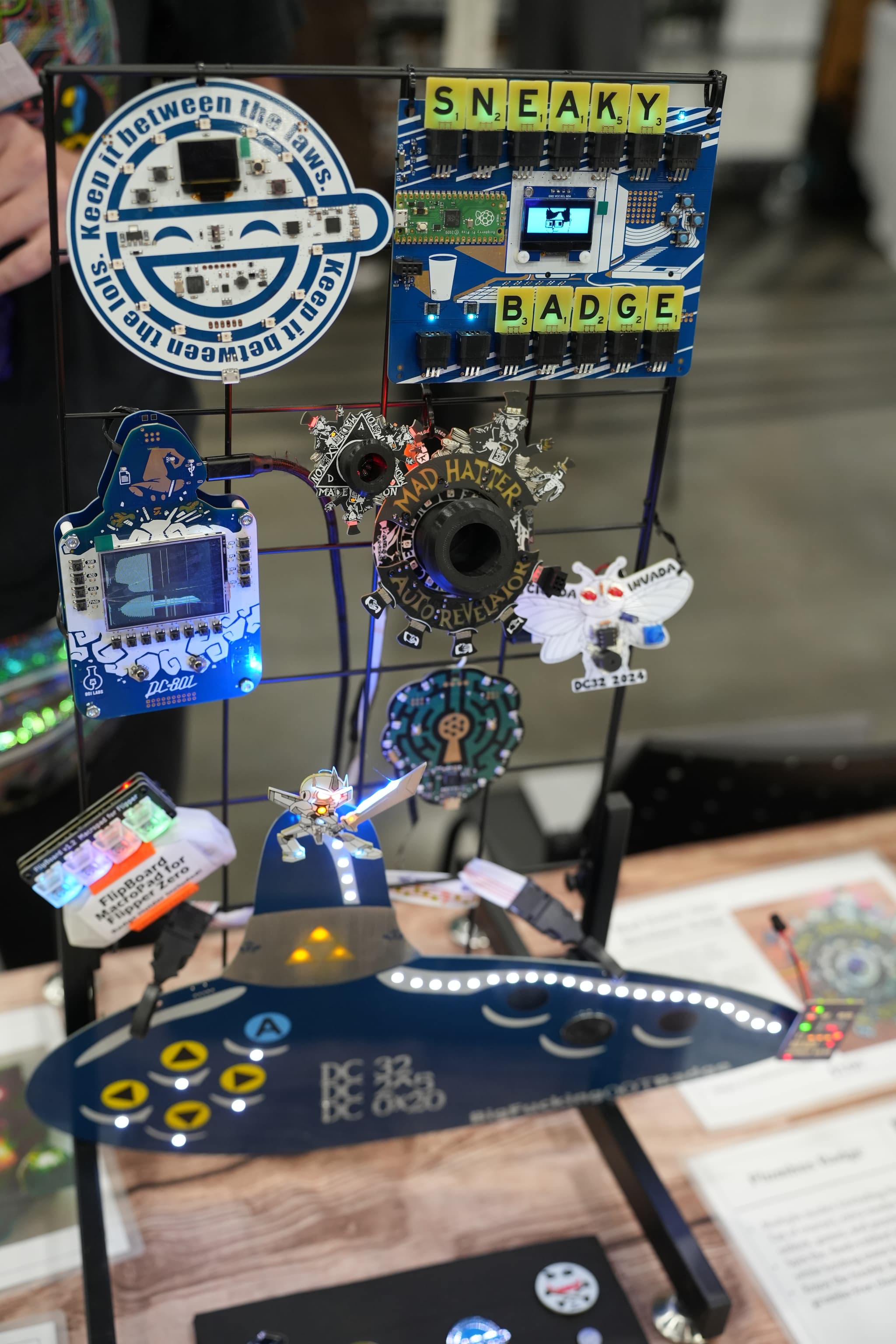 A display of various electronic badges and circuit boards, including one labeled SNEAKY BADGE, arranged on a metal stand