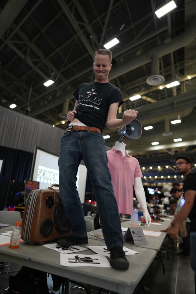 A person standing on a table, holding a belt and posing energetically in a large indoor venue with high ceilings and industrial lighting. There are various items on the table, including a suitcase and papers, and a mannequin with a pink shirt in the background