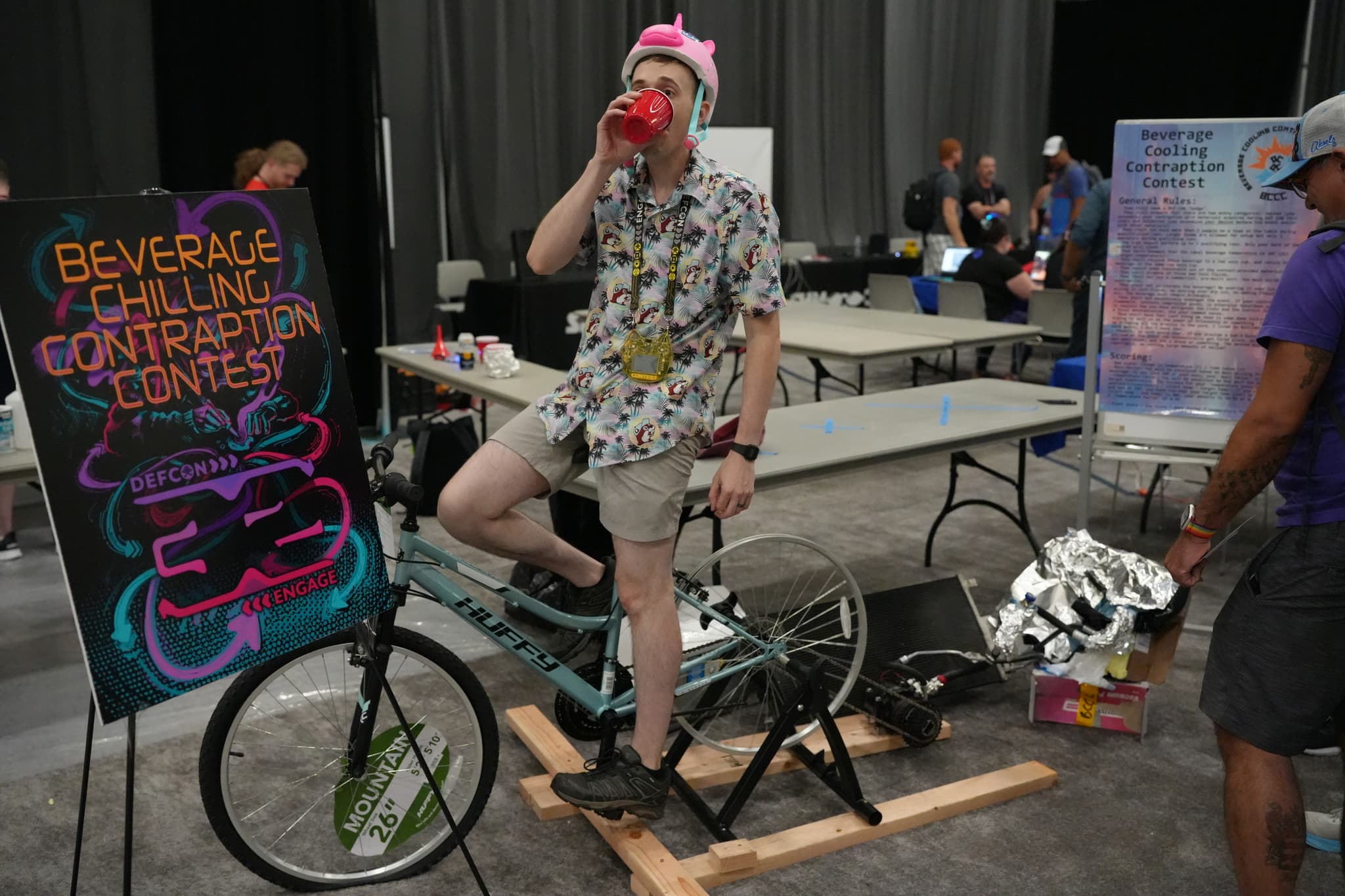 A person wearing a floral shirt and shorts is standing on a stationary bicycle, drinking from a cup. A sign next to them reads Beverage Cooling Contraption Contest. Several people are in the background, and tables with various items are set up around the area