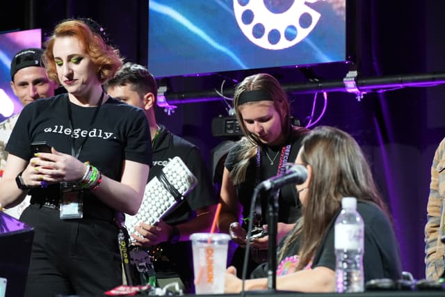 A group of people gathered around a table with microphones and drinks, some looking at their phones, in a setting with stage lighting and a large screen in the background