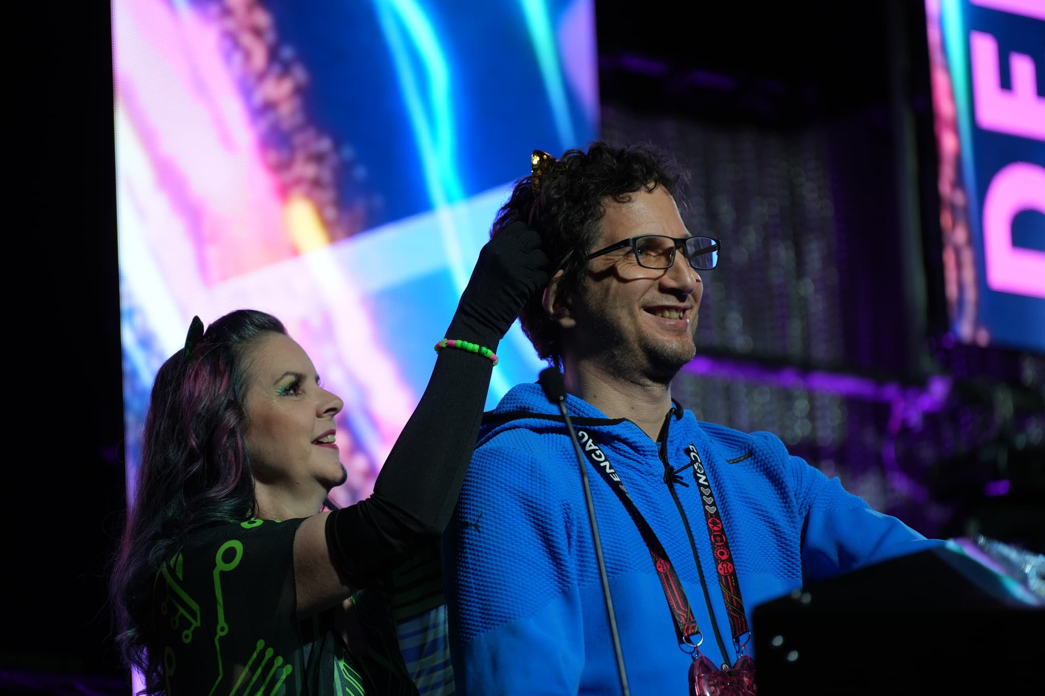 A woman in a black outfit and gloves is placing a crown on the head of a smiling man wearing glasses and a blue hoodie, with a colorful, illuminated background behind them