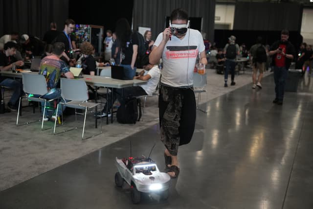 A person wearing a VR headset is controlling a small robotic vehicle with lights in an indoor setting, surrounded by people sitting at tables and engaging in various activities