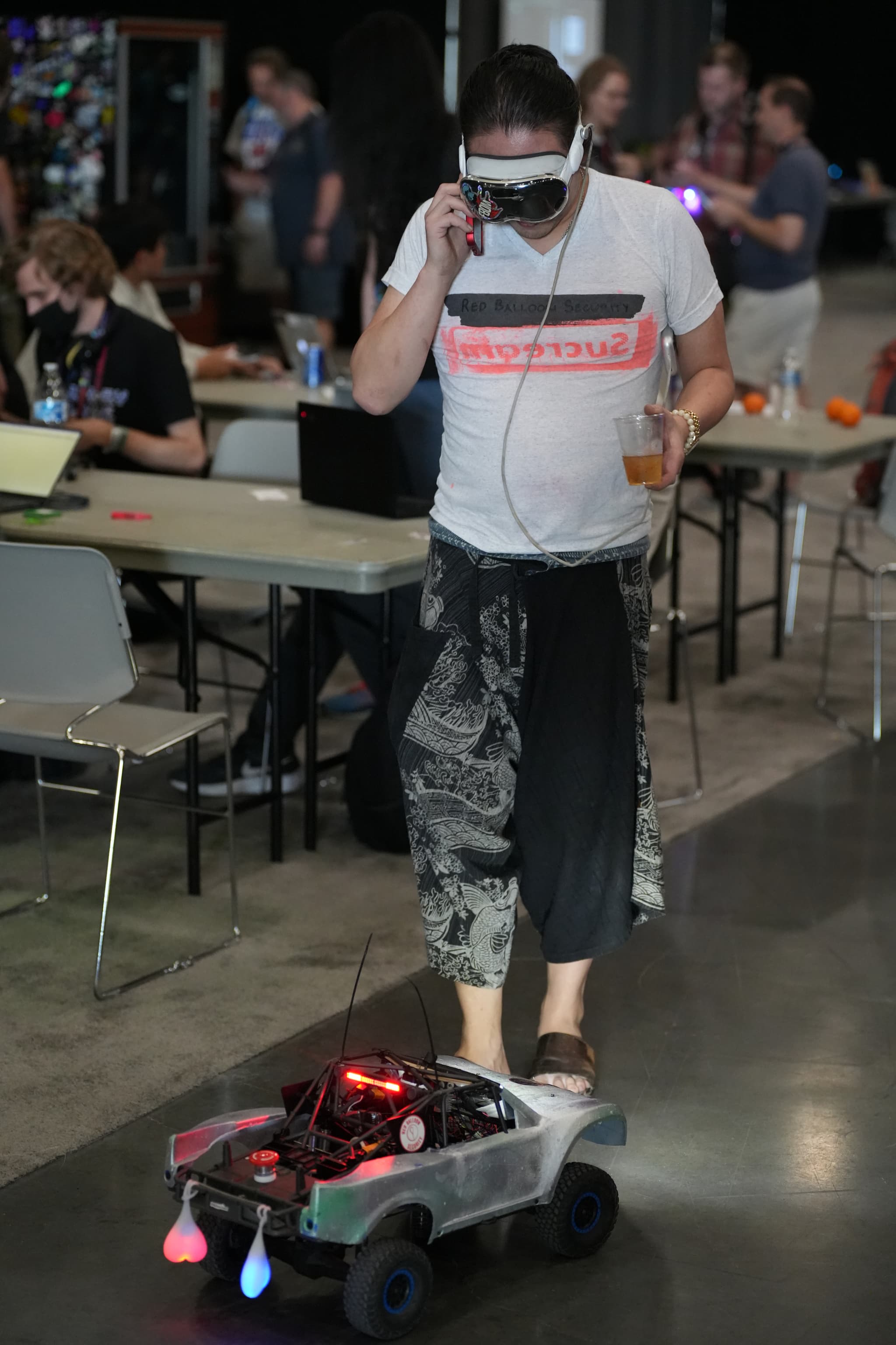 A person wearing a white t-shirt and black pants is controlling a small robotic vehicle with lights, using a remote control. The person is also wearing virtual reality goggles and holding a drink. In the background, there are tables with people working on laptops and various equipment