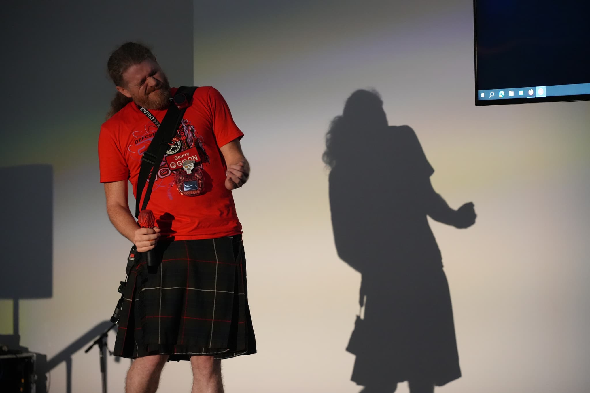 A person wearing a red shirt and a kilt, holding a microphone, with their shadow cast on a wall behind them