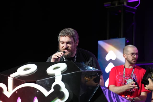 Two men are on a stage, one speaking into a microphone and the other holding a drink. The stage features a large, illuminated design with a stylized face