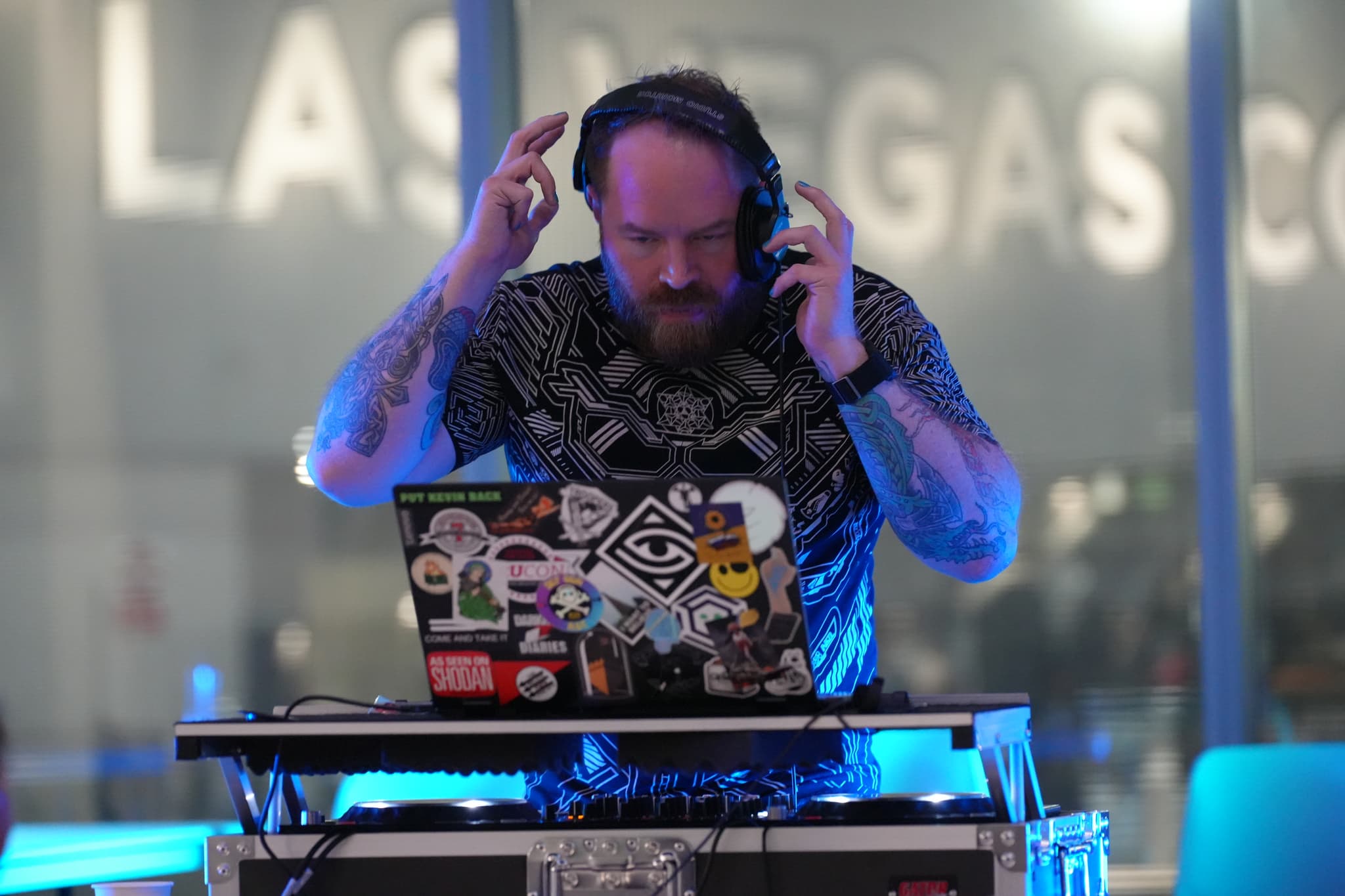 A DJ with tattoos is performing, using a laptop covered in stickers and a DJ controller. The background features large, blurred letters spelling LAS VEGAS