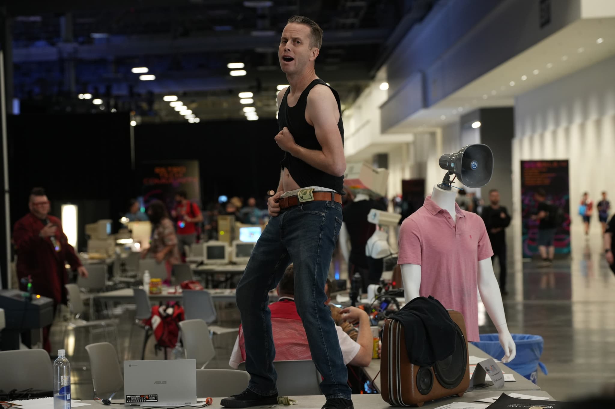 A person standing on a table in a large indoor space, wearing a black tank top and jeans, with a mannequin and various items around